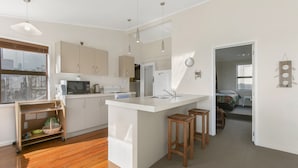A modern kitchen with a central island, wooden stools, and a view into the adjacent bedroom. - Sweeping Sea Views - Waihi Holiday Home (Waihi Beach)