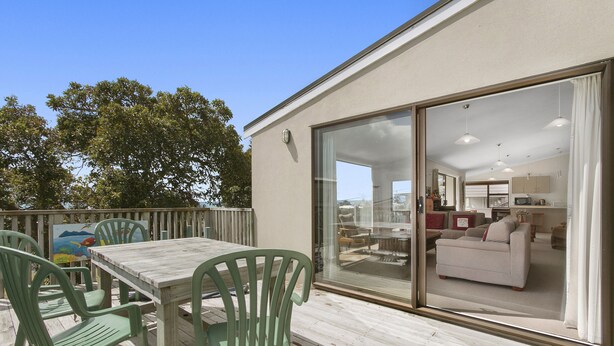 A deck with a table and chairs, a sliding glass door leading to a living area with a sofa and a dining table. - Sweeping Sea Views - Waihi Holiday Home (Waihi Beach)