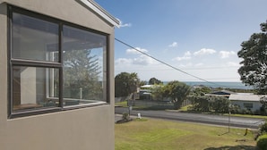 A residential building with large windows, a well-maintained lawn, and a view of a road and distant houses. - Sweeping Sea Views - Waihi Holiday Home (Waihi Beach)