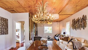 A wooden dining table with chairs, a chandelier, a sofa, and a bookshelf in a room with wooden ceilings and walls.