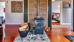 A wood-burning stove with logs, a stone fire surround, and a cozy living room with a bed and a painting of a deer.