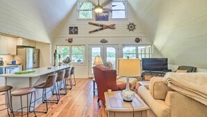 A spacious living area with a high ceiling, wooden walls, a kitchen with modern appliances, and a cozy seating area with a TV and lamp. - Lake Martin Cabin w/ Luxury Dock & Kayaks! (Eclectic)