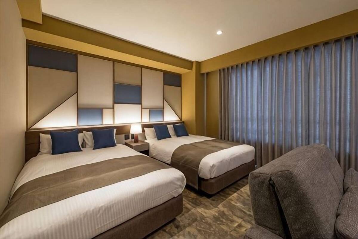 Habitación de hotel con dos camas, un sofá, una mesita de noche con lámpara y un ventanal con cortinas.