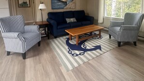 Un salon avec un canapé bleu, des fauteuils à rayures, une table basse en bois et un tableau sur le thème de la marine accroché au mur.