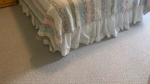Un lit bien fait, recouvert d’une couvre-lit à motifs floraux et agrémenté d’oreillers assortis, une table de chevet en bois avec une lampe, et un sol recouvert d’une moquette.