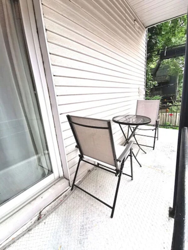 Un patio techado con una mesa metálica y dos sillas, un ventanal corredero y vista a un jardín trasero con una parrilla.