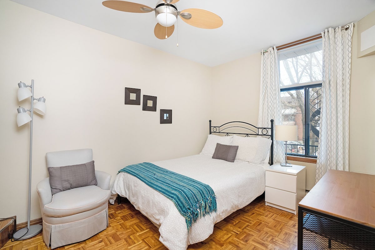 A bedroom with a bed, bedside table, chair, and a ceiling fan.