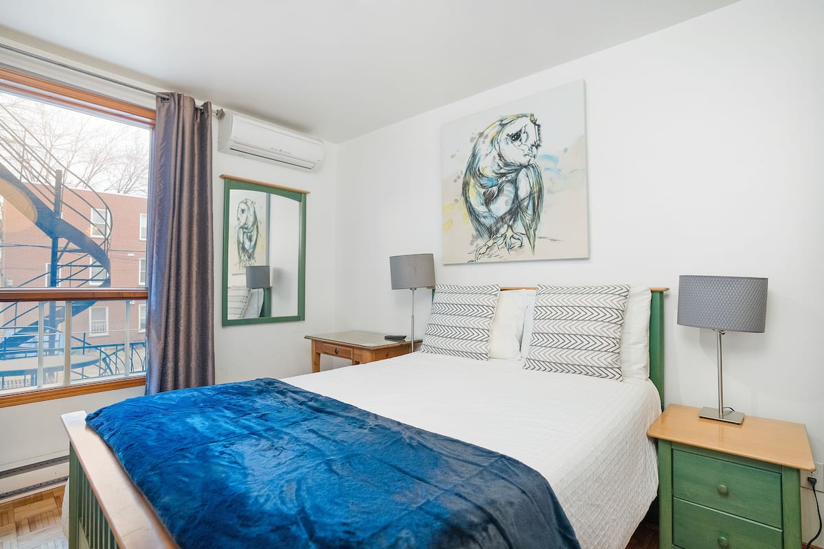 A bedroom with a bed, two bedside tables with lamps, a wall-mounted painting, and a window with a view of buildings.