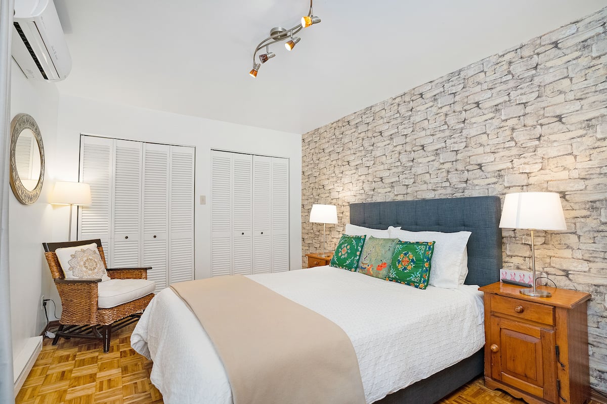 A bedroom with a large bed, two bedside tables with lamps, a wicker chair, a stone wall, and a wardrobe with white shutters.