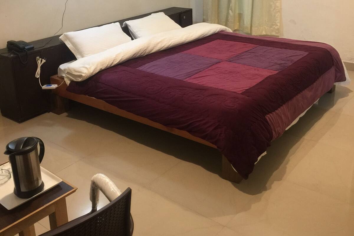 Una cama bien hecha con una colcha rojo oscuro, una mesita de noche con una lámpara y un enchufe en la pared.