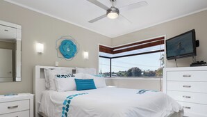 Un dormitorio con cama, mesita de noche, cómoda, ventilador de techo, ventana con persianas y televisor en la pared.