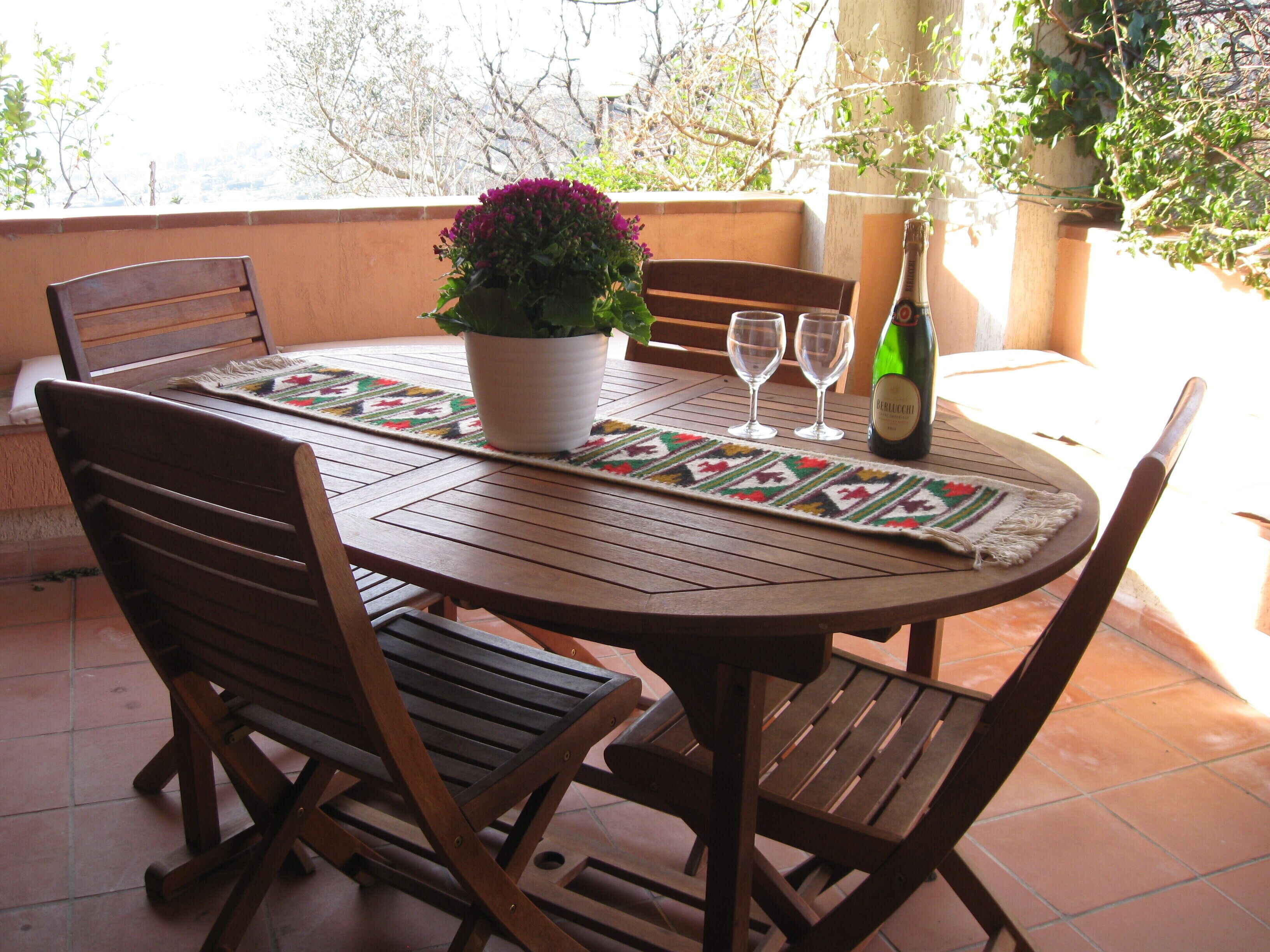 Una mesa de madera redonda con un posavasos tejido, dos copas de vino, una botella de vino y una planta en maceta con flores moradas.