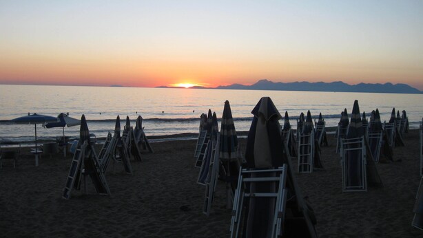 A beach with rows of sun loungers and umbrellas, a sunset in the background, and mountains in the distance.