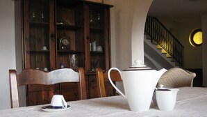 A dining table with a white tablecloth, a white teapot, a white cup, and a saucer. There is a wooden chair with a white cushion. In the background, there is a wooden cabinet with glass doors containing various items.