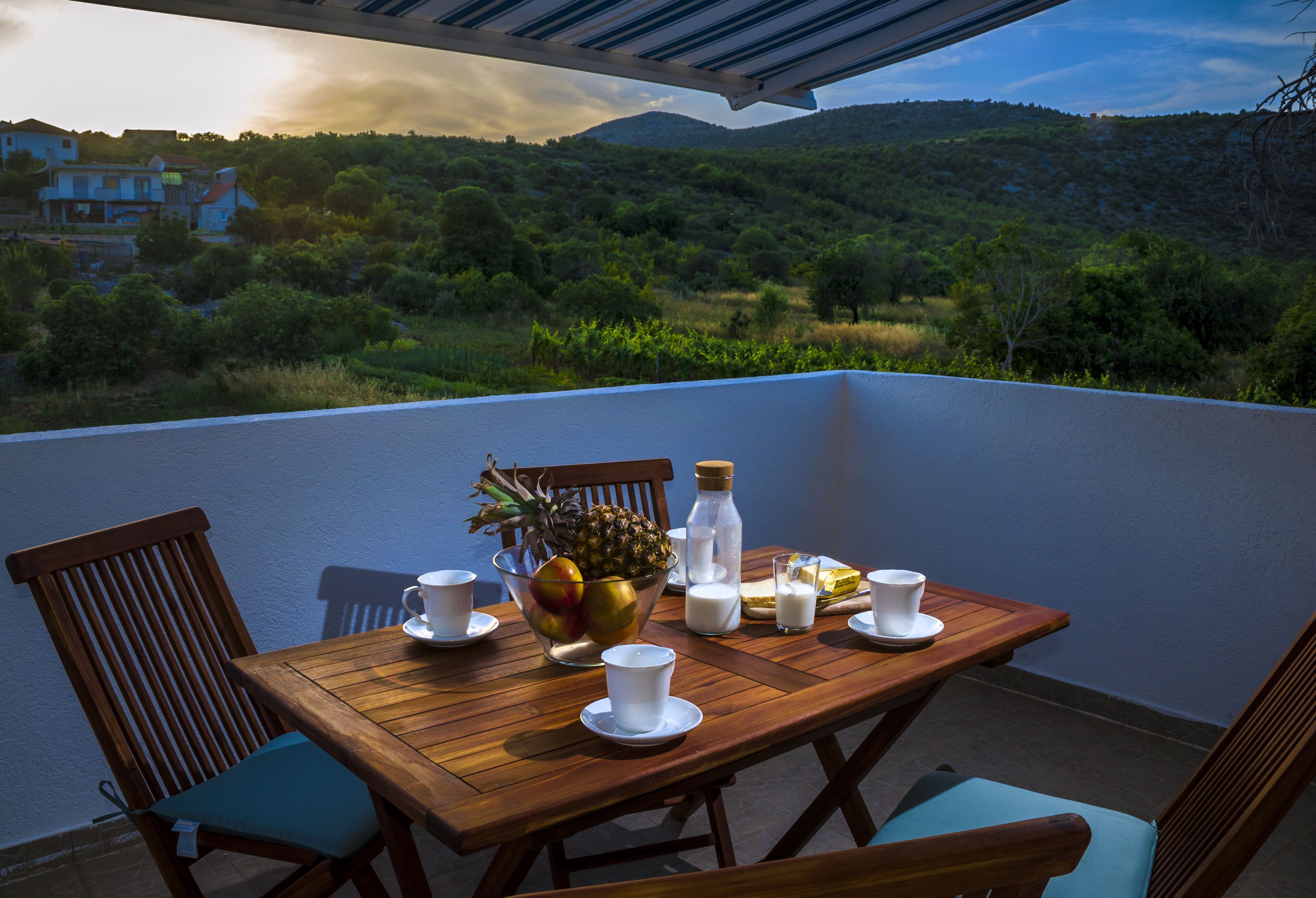 A wooden table with a bowl of fruit, milk, and cups on a balcony overlooking a hillside.