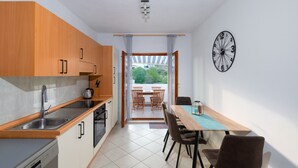 A modern kitchen with wooden cabinets, a stainless steel sink, and a dishwasher. There is a dining area with a table and chairs, and a large clock on the wall. - Enjoy your private pool and outdoor kitchen (Marina)
