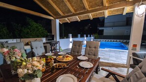 An outdoor dining area with a wooden table set for a meal, surrounded by chairs, and a view of a swimming pool in the background. - Enjoy your private pool and outdoor kitchen (Marina)