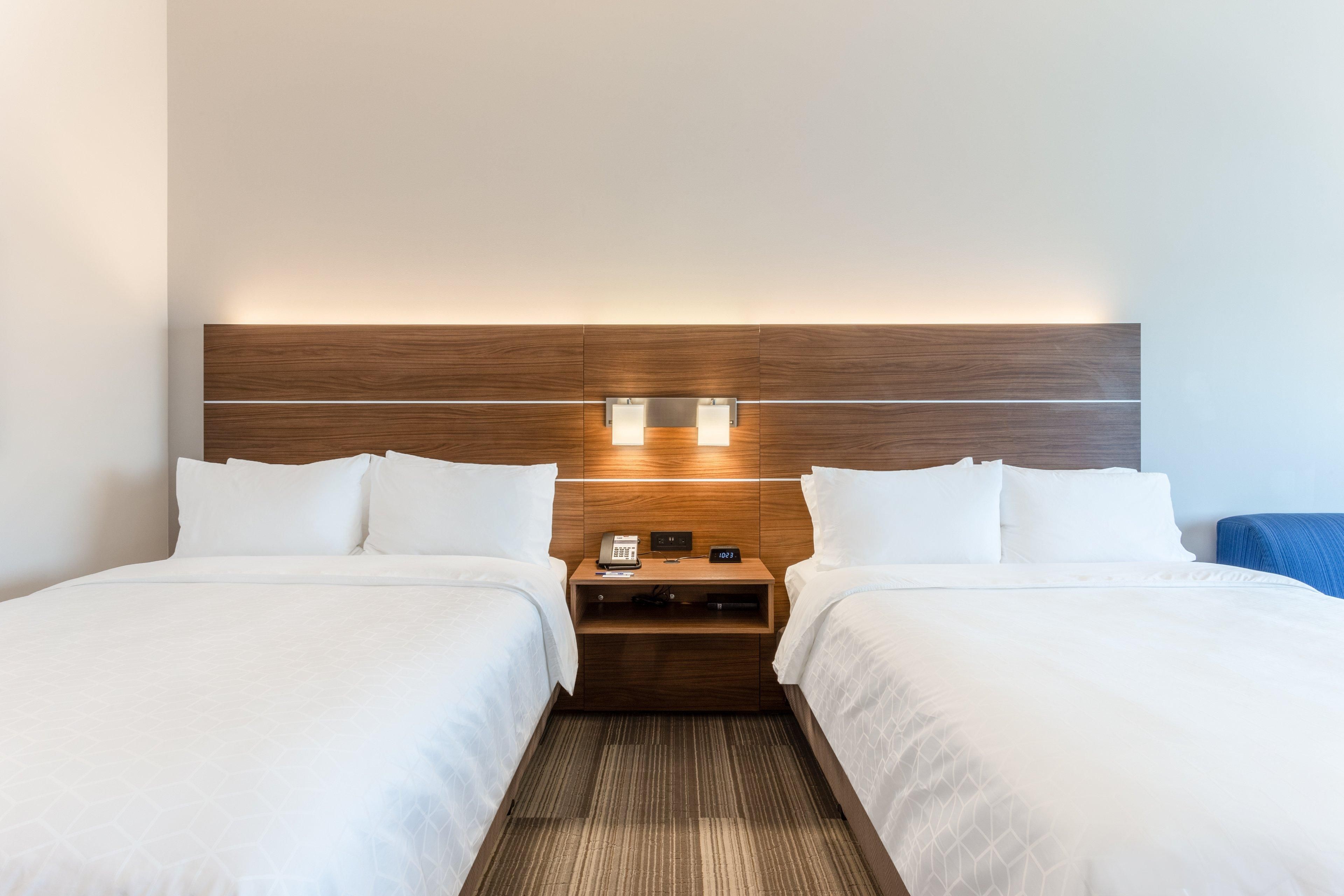 A hotel room with two beds, a wooden headboard, bedside tables with lamps, and a small nightstand with a phone and a lamp.