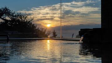 A swimming pool with a sunset in the background, a water feature on the right, and trees around the pool.