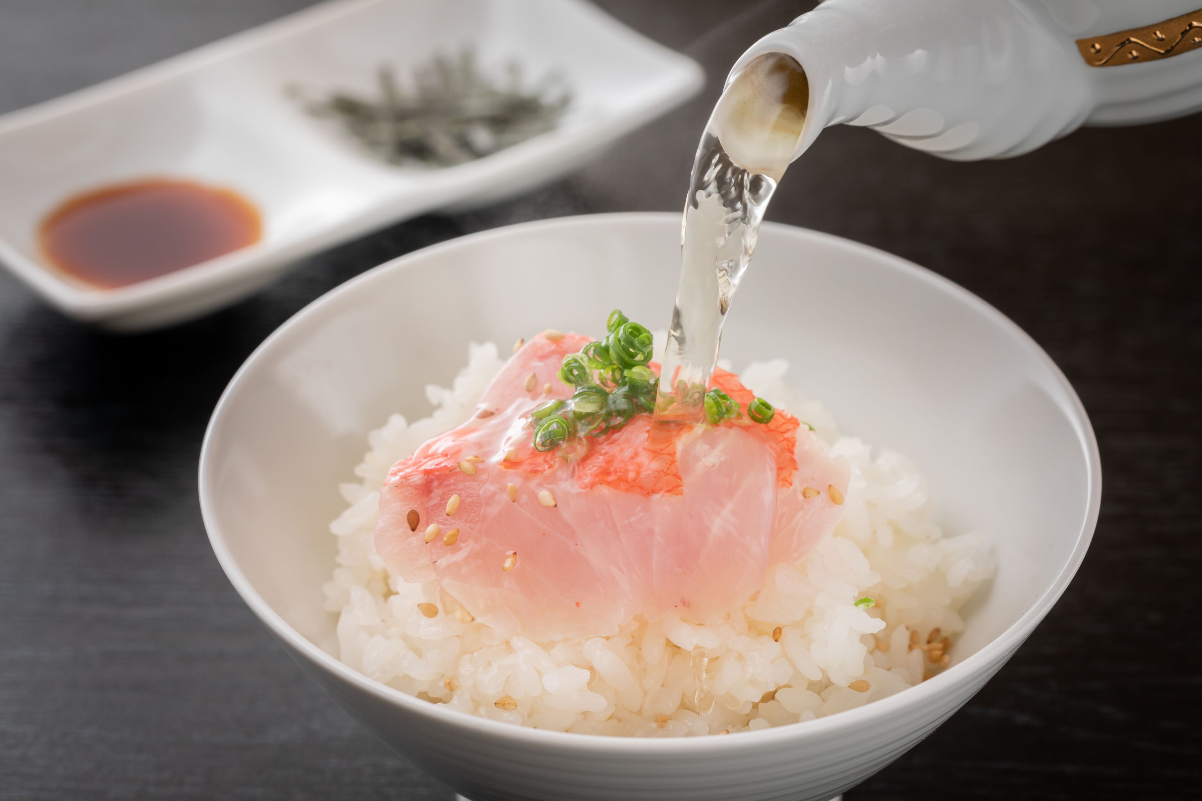A bowl of sushi rice topped with salmon and green onions, with a small dish of soy sauce in the background.