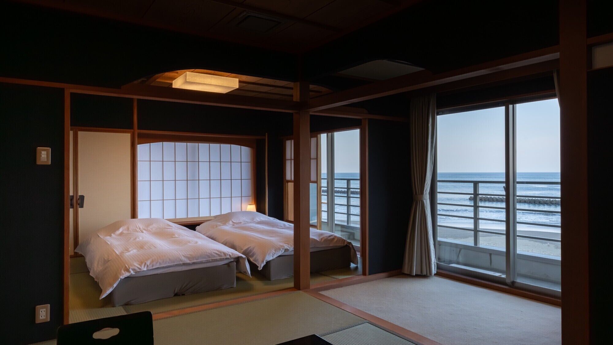 Una tradizionale stanza in stile giapponese con pavimento in tatami, un letto basso e uno schermo shoji, e vista sul mare attraverso ampie finestre.