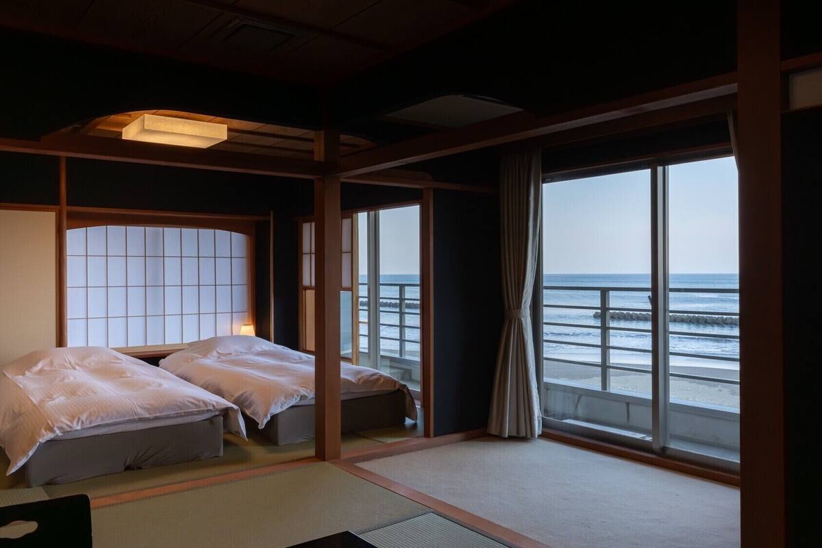 A traditional Japanese-style room with tatami flooring, a low bed with a shoji screen, and a view of the sea through large windows.