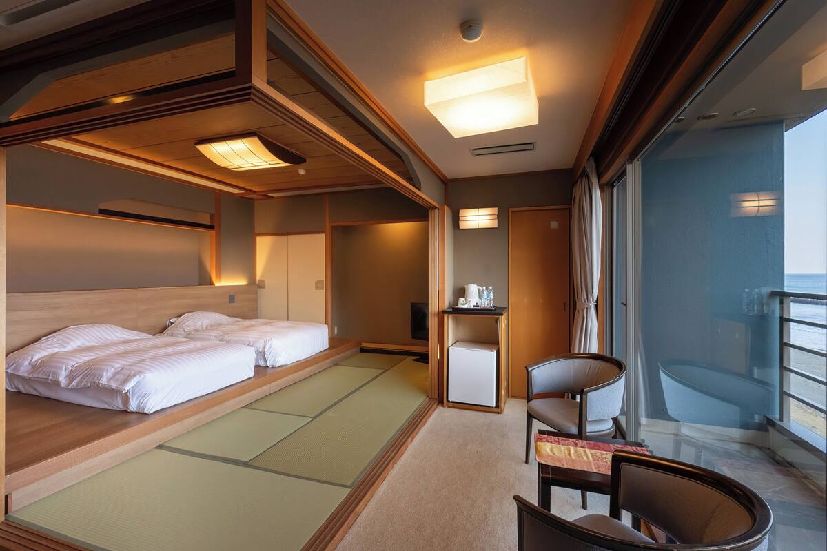 A hotel room with a bed, a small table with a kettle, a chair, and a view of the sea.