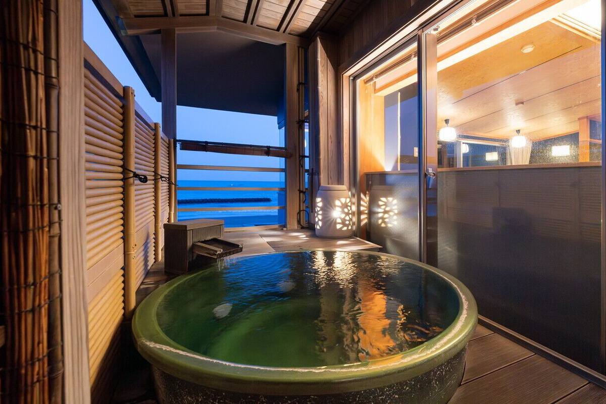 A hot tub with a view of the ocean, a wooden deck, and a glass door leading to a room.