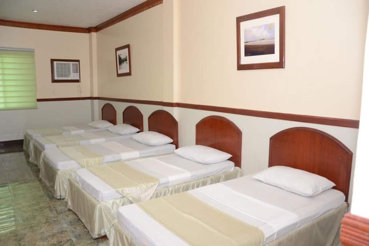 Una habitación de hotel con cuatro camas individuales, cada una con cabecero de madera y colcha color beige. Hay un cuadro enmarcado en la pared sobre las camas, y un equipo de aire acondicionado montado en la pared a la izquierda.