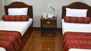 Habitación de hotel con dos camas individuales, cada una con cabecero de madera, ropa de cama blanca y almohadas estampadas en rojo. Entre las camas hay una mesita de noche con un teléfono, una lámpara y un cartel. El piso es de baldosas.