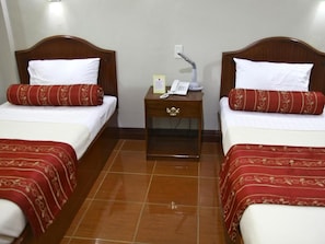A hotel room with two single beds, each with a wooden headboard, white bedding, and red patterned pillows. There is a nightstand between the beds with a telephone, a lamp, and a sign. The room has a tiled floor. - Villa Maria Luisa Hotel (Tandag)