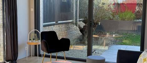 A modern living room with a sofa, a coffee table, and a chair. There is a large sliding glass door leading to an outdoor area with plants and a red wall.