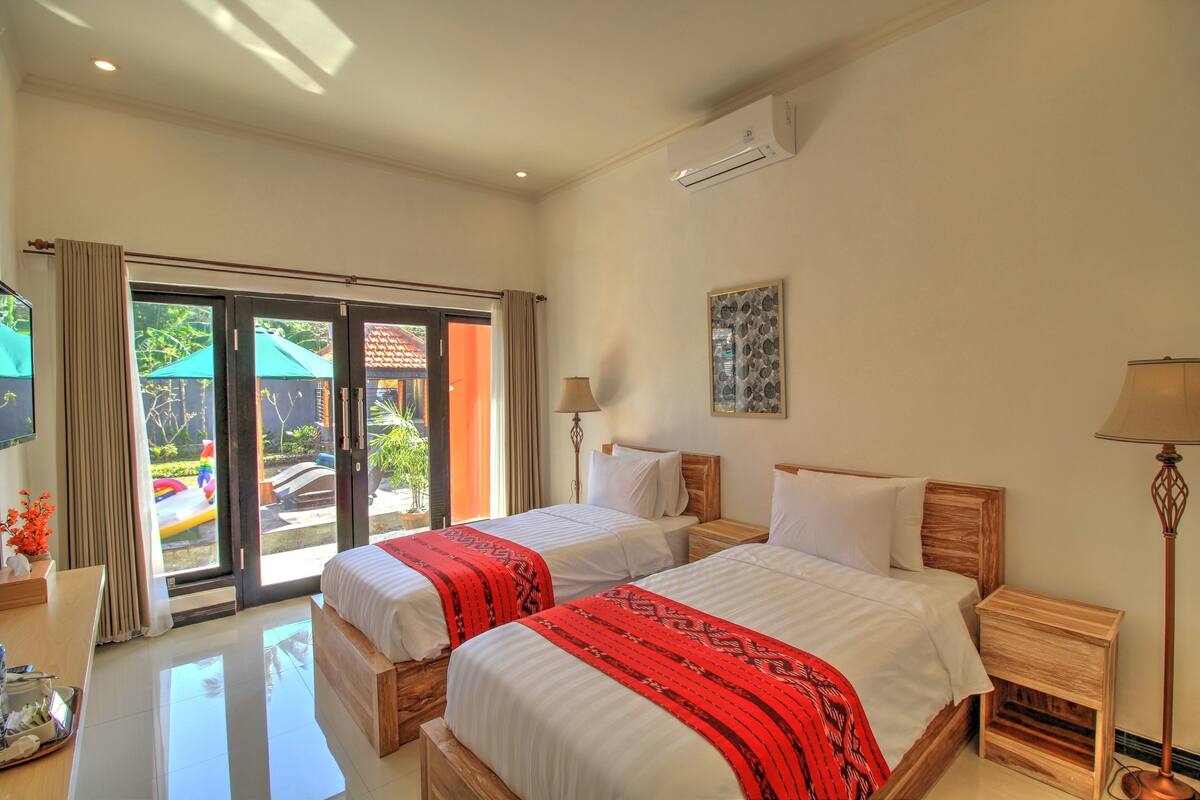 A hotel room with two beds, a desk, a lamp, and a view of the outside.