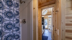 Un couloir intérieur en bois, avec un rideau de douche à motifs, une porte donnant sur un coin cuisine équipé de meubles en bois et d’un four à micro-ondes, et une horloge visible au mur.