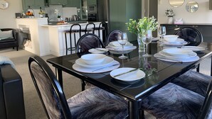 A modern dining area with a glass table set for six, surrounded by dark upholstered chairs. A kitchen with green cabinets and a white countertop is visible in the background.