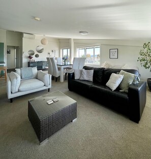 A living room with a white sofa, a black leather sofa, a coffee table, and a dining area with chairs and a table.