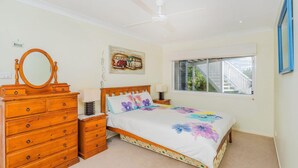 Un dormitorio con cama, cómoda de madera, espejo, letrero en la pared, ventana y ventilador de techo.