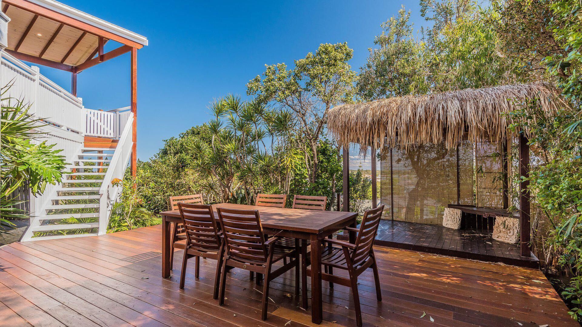 A wooden deck with a dining table and chairs, surrounded by lush greenery and a thatched-roof structure.