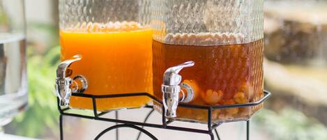 Two glass dispensers with labels for orange and longan juice, placed on a table with a view of greenery outside.