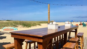 A wooden deck with a table and benches, a sandbar sign, and beach umbrellas in the background.
