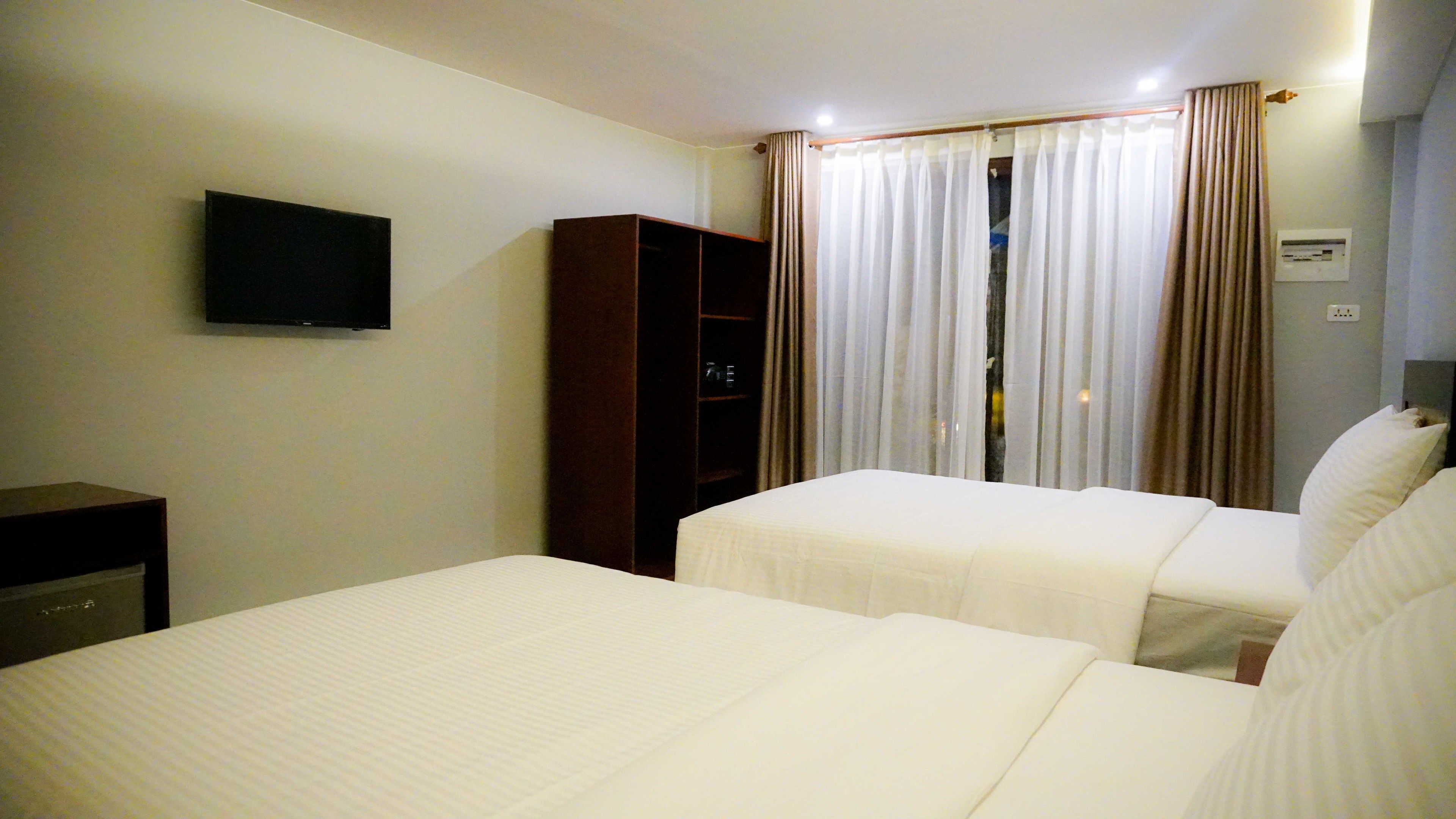 Ein Hotelzimmer mit zwei Betten, einem an der Wand befestigten Fernseher, einem Kleiderschrank und einem Fenster mit Vorhängen.