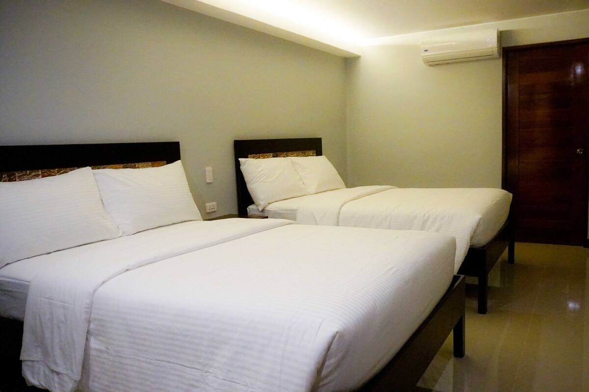 Habitación de hotel con dos camas individuales, cabecero de madera, escritorio y unidad de aire acondicionado en la pared.