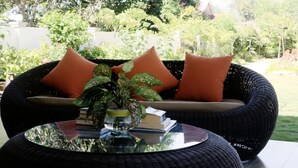 Ein Korbsessel mit orangefarbenen Kissen, ein Glastisch mit Pflanze und Büchern sowie Blick auf einen Garten.