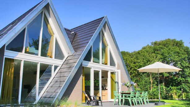 Ein modernes Haus mit großen Fenstern, einem geneigten Dach und einer Terrasse mit Gartenmöbeln.