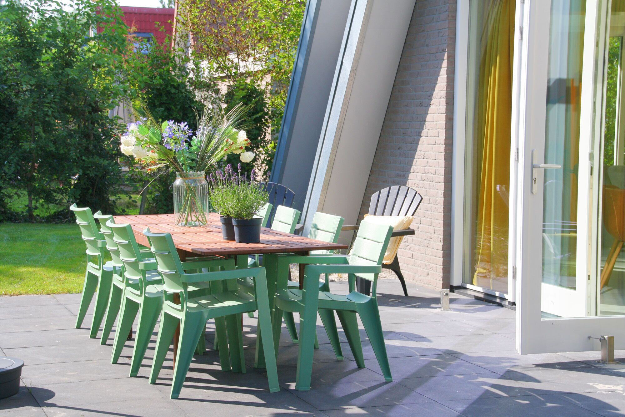 Un patio al aire libre con una mesa de madera y sillas verdes, rodeado de plantas en macetas y un ventanal que da a un edificio.
