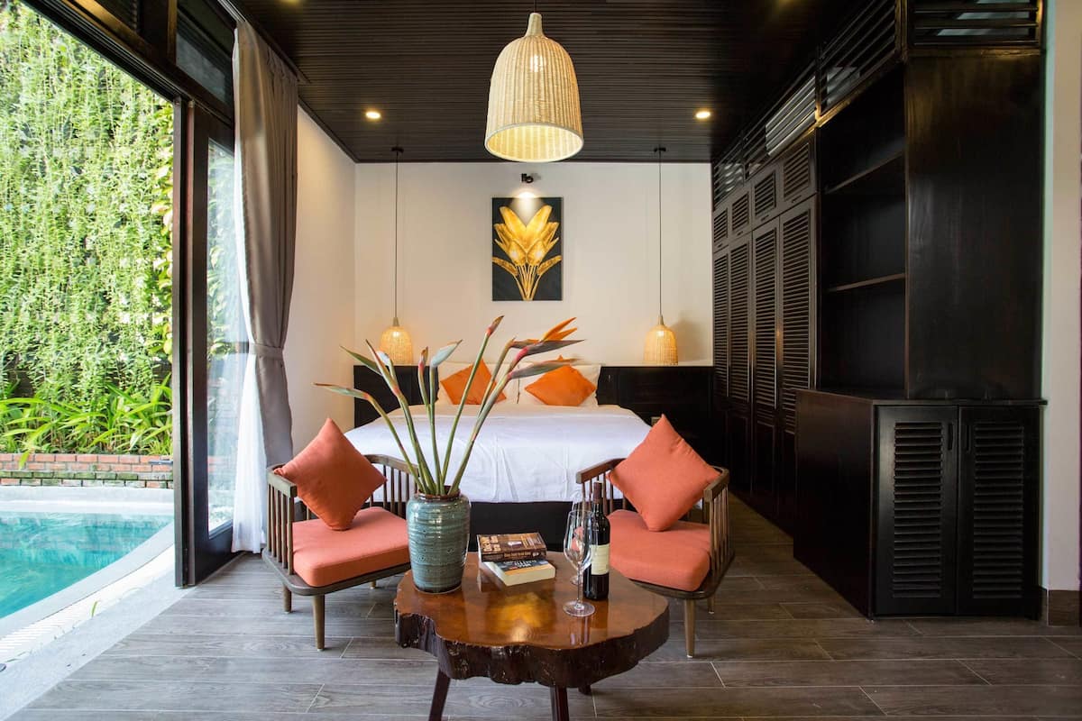 Ein Schlafzimmer mit einem Bett, zwei Stühlen, einem Tisch mit einer Vase und Büchern, einem Weinglas und einem großen Fenster mit Vorhängen.