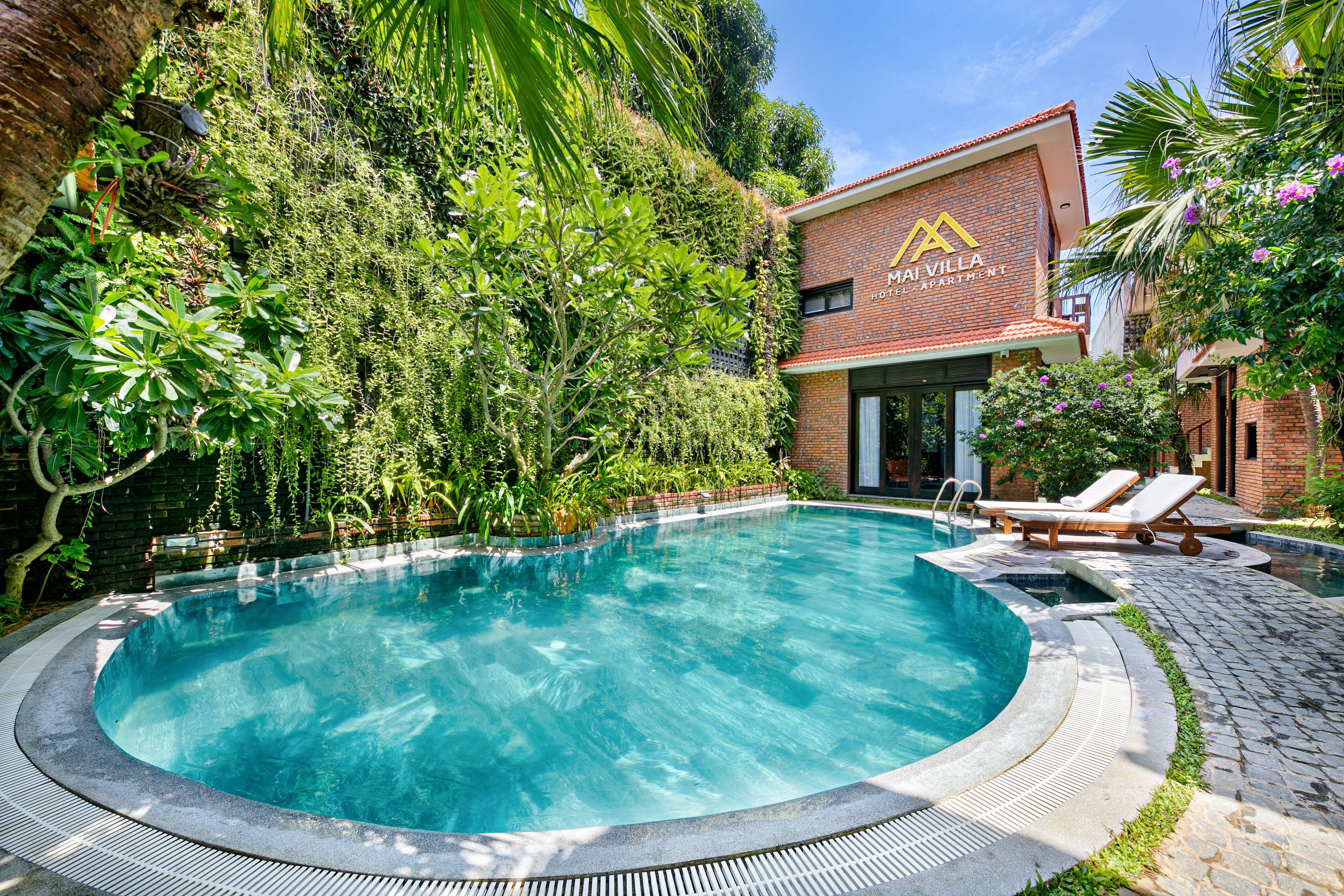 A swimming pool surrounded by lush greenery and a building with a sign reading "MAI VILLA HOTEL APARTMENT".