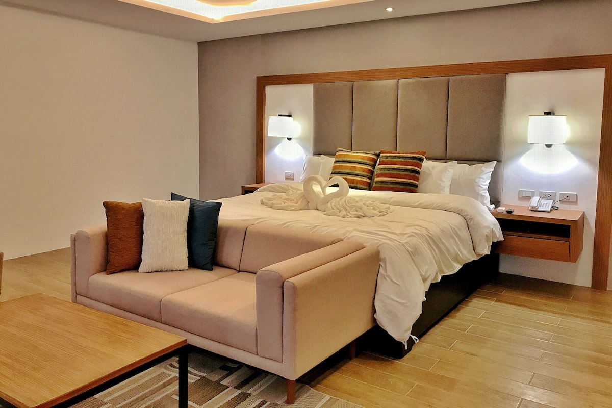 Una habitación de hotel moderna con una cama grande, un sofá, una mesita de noche con lámpara y suelo de madera.