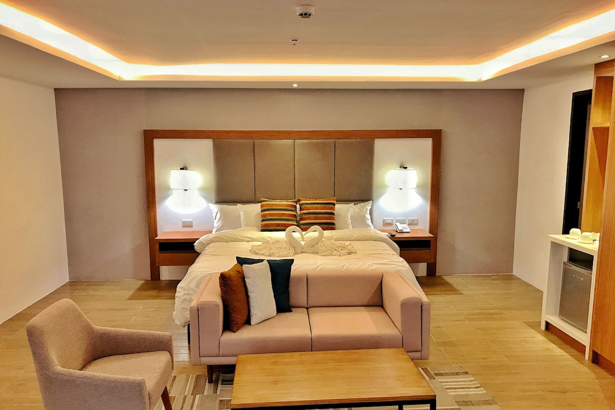 Una habitación de hotel moderna con una cama grande, un sofá, una silla y una mesita. Hay dos lámparas de noche y un espejo sobre la cama.
