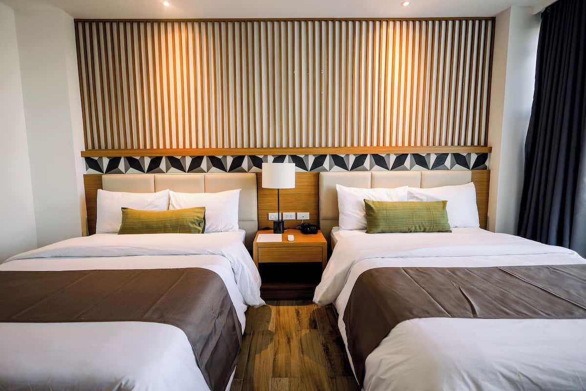 Habitación de hotel con dos camas, cabecero de madera, mesita de noche con lámpara y pared con estampado geométrico.